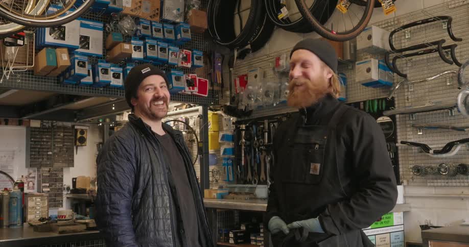The interior of a bicycle repair shop with two individuals engaged in conversation amidst neatly arranged bicycle parts and tools on shelves and walls