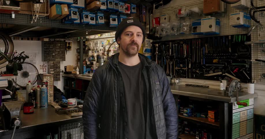A man in a black jacket and wearing a cap is standing in a workshop and looking straight ahead