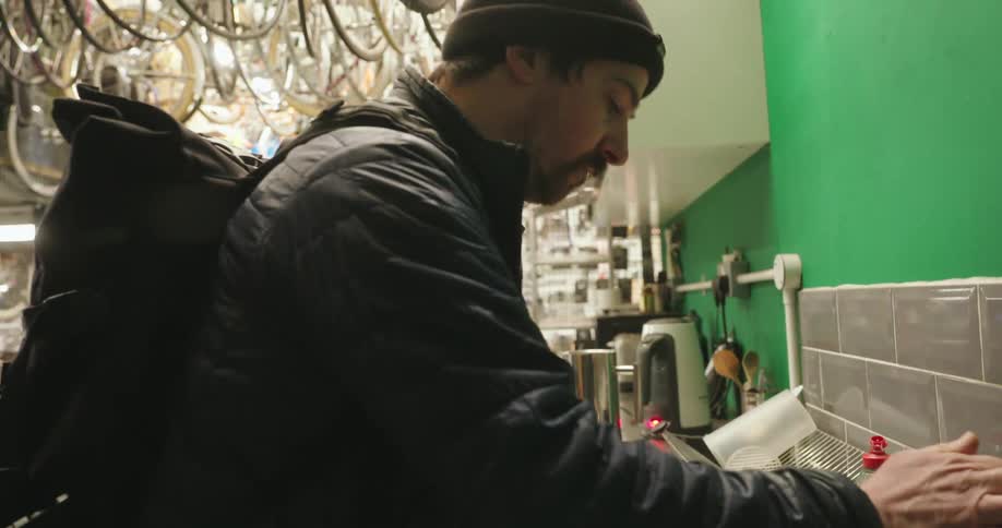 A man stands in a bicycle shop and fills a metal coffee carafe with water