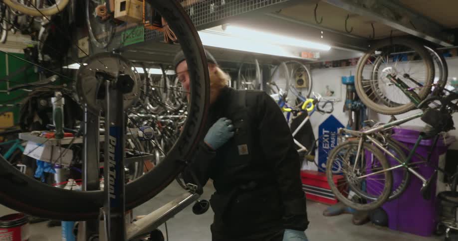 A cluttered workshop with a person repairing a bicycle surrounded by scattered tools and spare parts