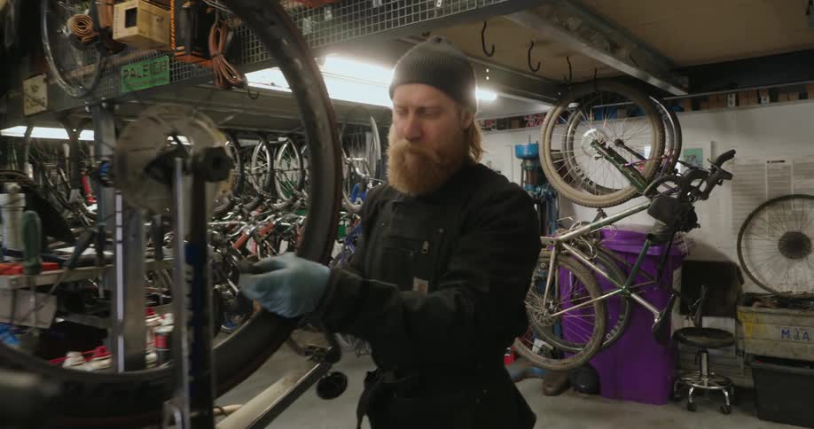 A bearded mechanic is carefully aligning a bicycle wheel on a repair stand in a cluttered bike workshop