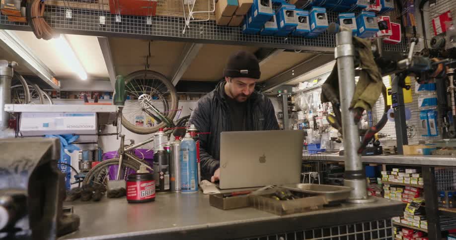 A man wearing a beanie stands at a cluttered workbench in a bicycle repair shop using a laptop computer