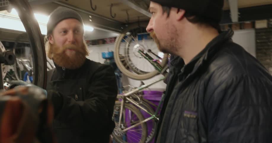 A bearded mechanic is intently adjusting a bicycle wheel when a man approaches for a conversation within the cluttered workshop