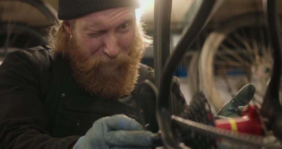 A bearded man in a beanie and gloves squints as he works intently on the rear gears of a bicycle in a workshop