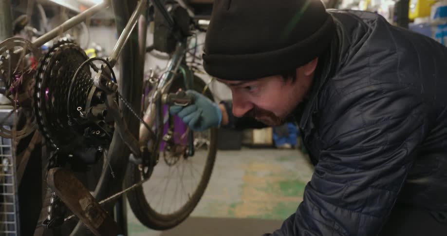 A person works on repairing a bicycle in a workshop filled with tools spare parts and hanging bikes