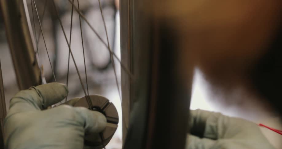 Gloved hands use a specialized spoke wrench tool to carefully adjust the tension of spokes on a bicycle wheel in a closeup view