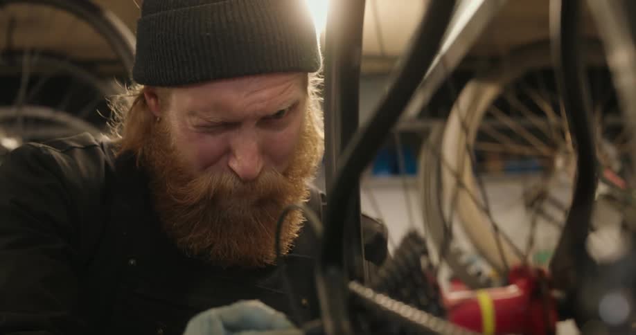 A bearded man in a beanie intently spins a bicycle wheel using his thumb as a reference point to check its alignment in a workshop