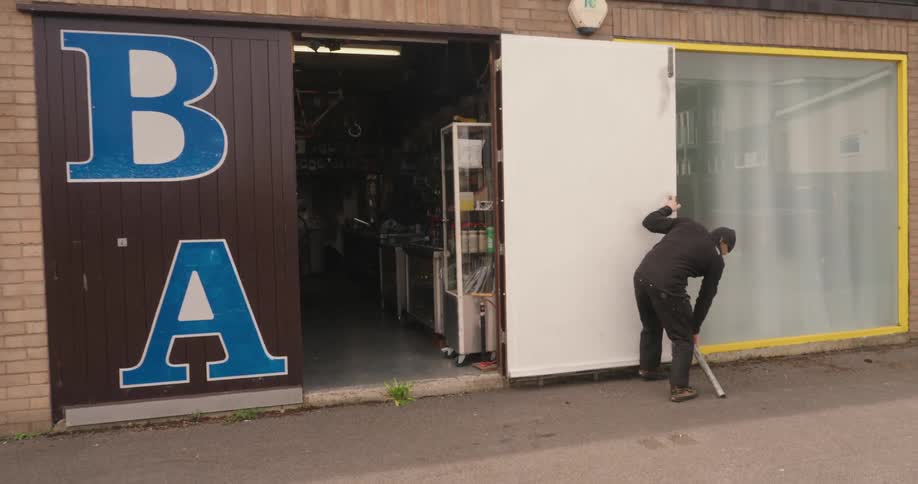 A bearded man slides open the large b a marked door of a workshop and props it open with a metal pole