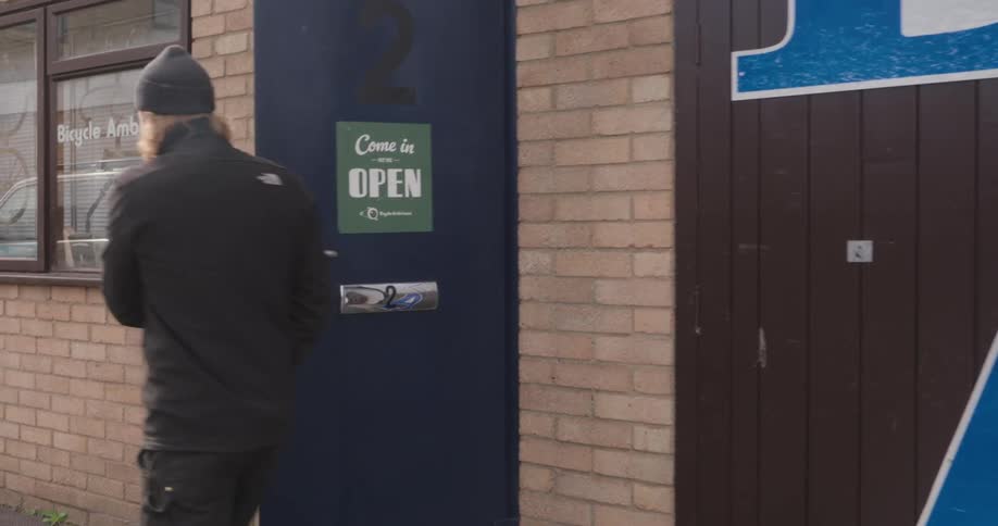 A man emerges from a blue door labeled 2 and places a green come in were open sign for bicycle ambulance onto it before walking away
