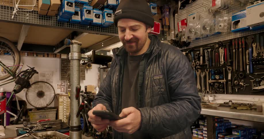 A man wearing a beanie and puffer jacket is texting on his smartphone amidst the tools and bicycles of a cluttered repair shop