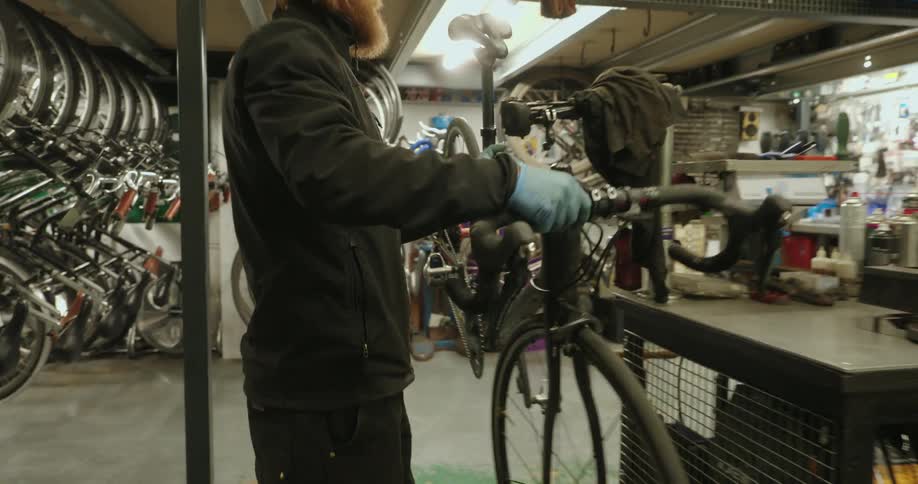 A bearded bike mechanic wearing a beanie lifts a bicycle and mounts it securely onto a repair stand in his workshop before starting work on it