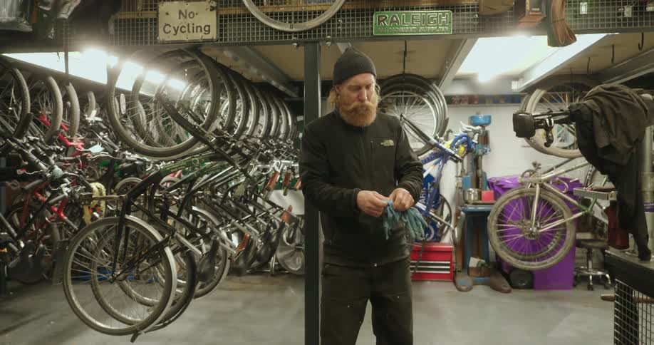Surrounded by numerous bicycles in a workshop a bearded man puts on work gloves and approaches a bike mounted on a repair stand