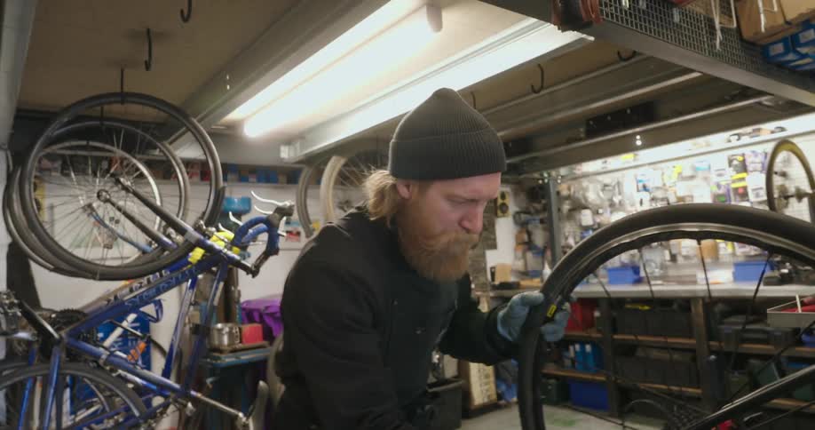 A bearded mechanic wearing gloves works on repairing a bicycle wheel and gears in a cluttered bike shop