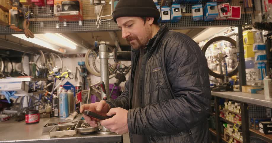 Man in cap and leather jacket standing in a bicycle workshop interacting with a smartphone