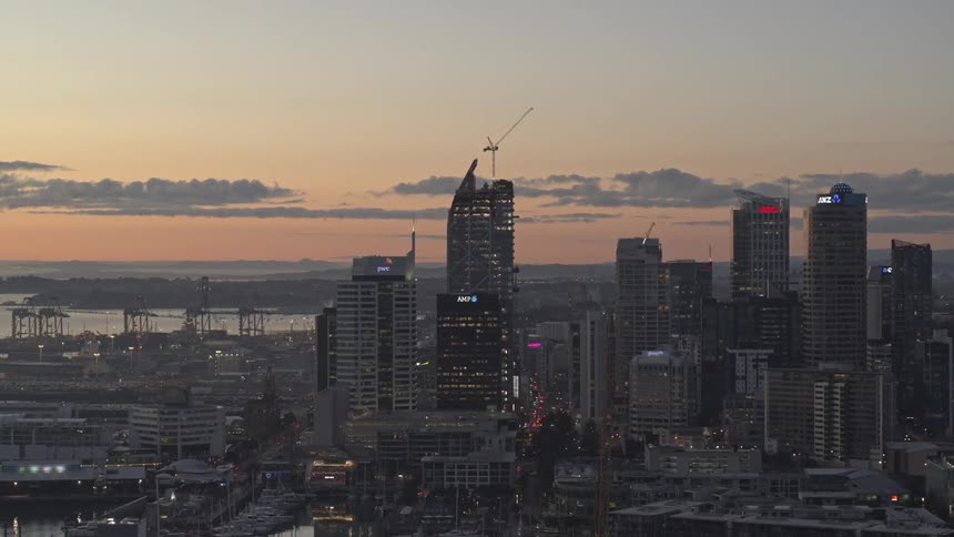A cityscape at dusk showcasing towering skyscrapers cranes and a harbor under a clear sky transitioning from orange to blue