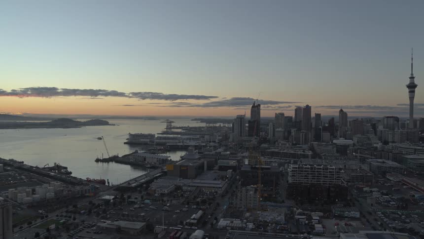 A panoramic view of auckland at sunrise showcases the skyline with the sky tower surrounding buildings and boats docked in the harbor