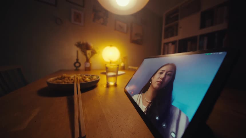 A person has a video call on a tablet while eating noodles at a dimly lit dining table