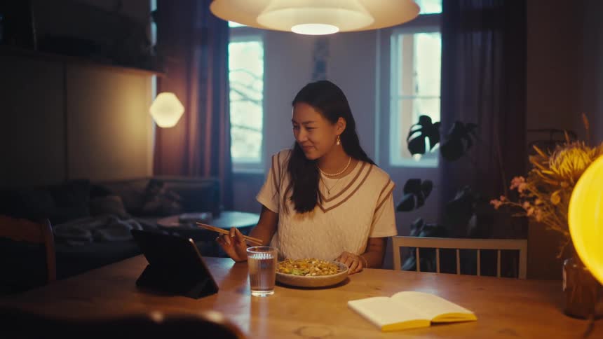 A person has a video call on a tablet while eating noodles at a dining table in a dimly lit cozy room