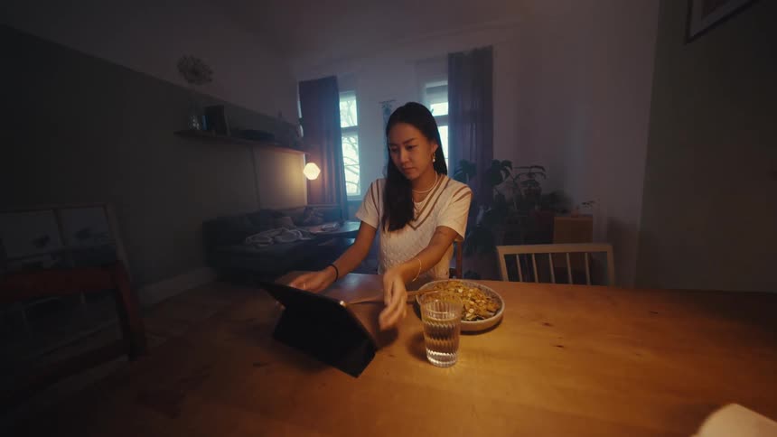 A dynamic zoom focuses on an asian woman as she sits at a dining table places a tablet down and looks at its screen with food and water nearby