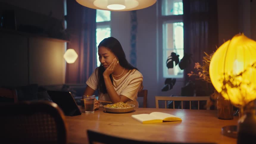 An asian woman sits at a dining table video chatting on a tablet holding chopsticks over her meal in a warmly lit room