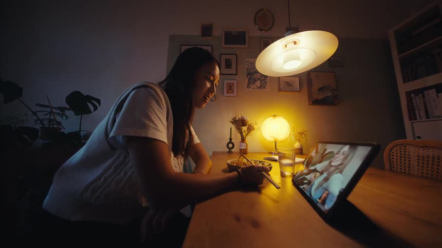 A person eats noodles from a bowl while using a tablet for a video call seated at a wooden dining table in a softly lit room with framed pictures on the walls