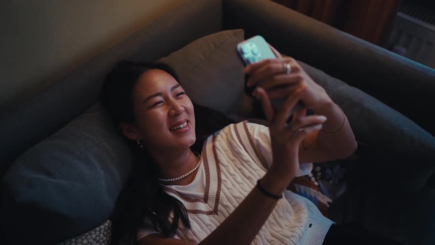 A young woman smiles and laughs while looking at her smartphone as she relaxes lying on a couch in a dimly lit room