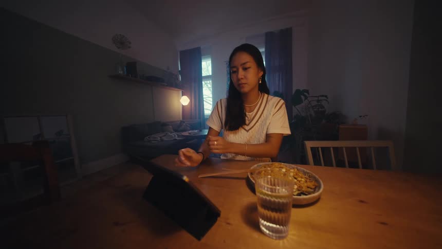 A woman sits at a wooden table with a bowl of food a glass of water and a tablet in a dimly lit room with large windows