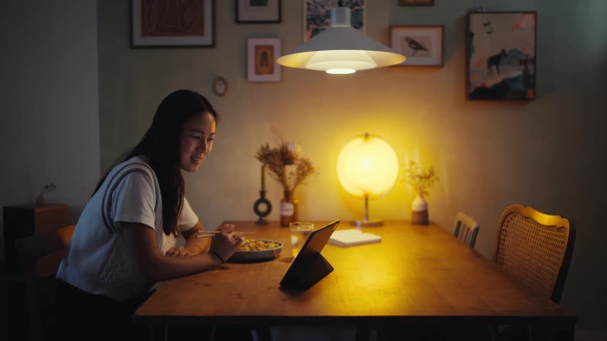 A young woman eating at the dining table while facetiming on a tablet placed on a stand a sphere light brightens the atmosphere with picture frames hanging on the wall
