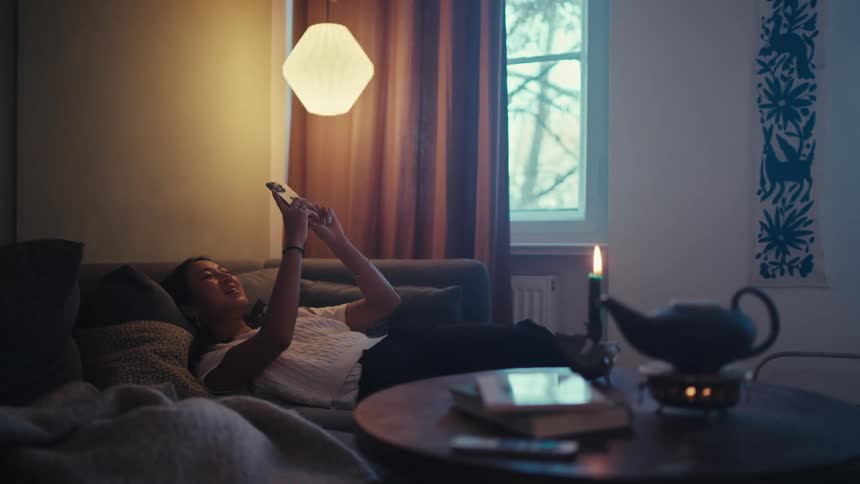 A young woman reclines on a couch in a dim room holding a phone with a candle and teapot on a nearby table and tree branches visible through the window