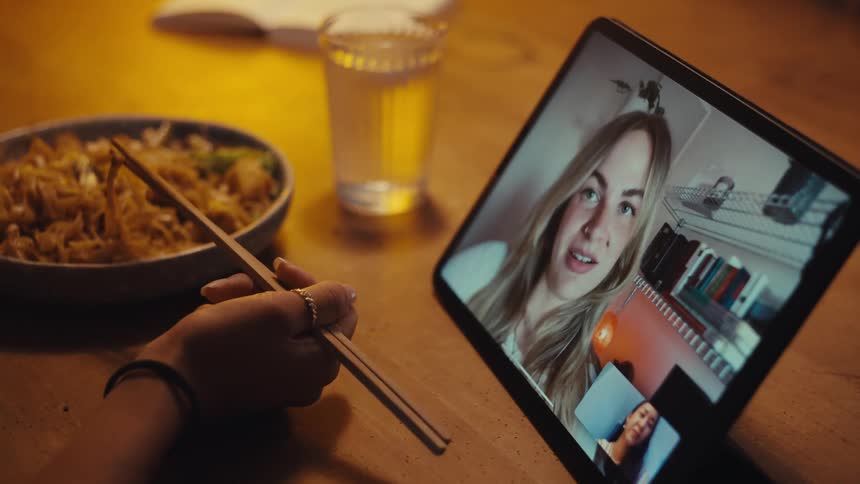 A person enjoys noodles using chopsticks while engaging in a video call on a tablet with a glass of water and an open book on the table