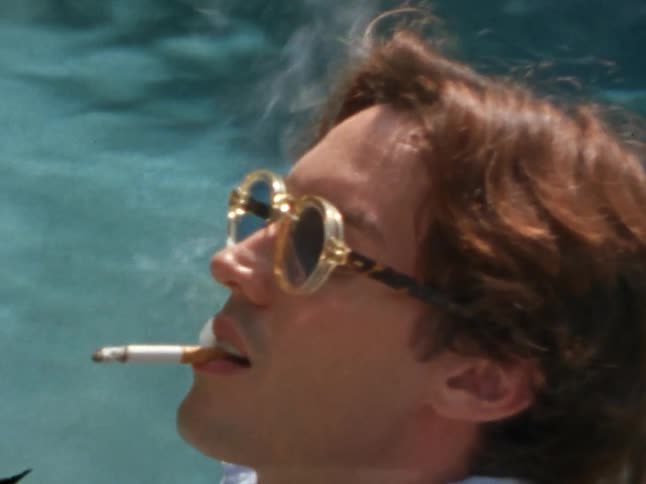 A person with brown hair and fashionable sunglasses smokes a cigarette beside a swimming pool
