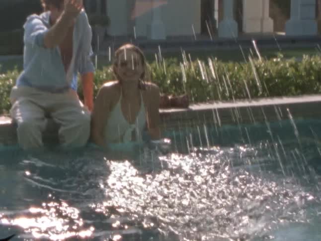 A young stylish couple sits on the pools edge playfully splashing water under the bright morning sun
