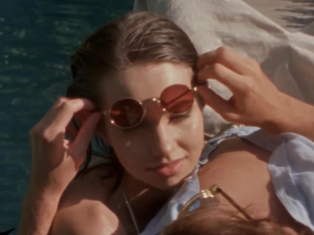 Woman at the edge of a pool leans against a man as he raises her sunglasses on a summer day