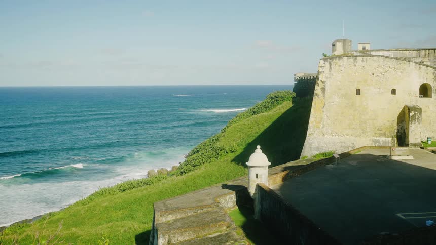 A historic stone fortress with watchtowers stands on a grassy cliff overlooking the ocean under a clear sky