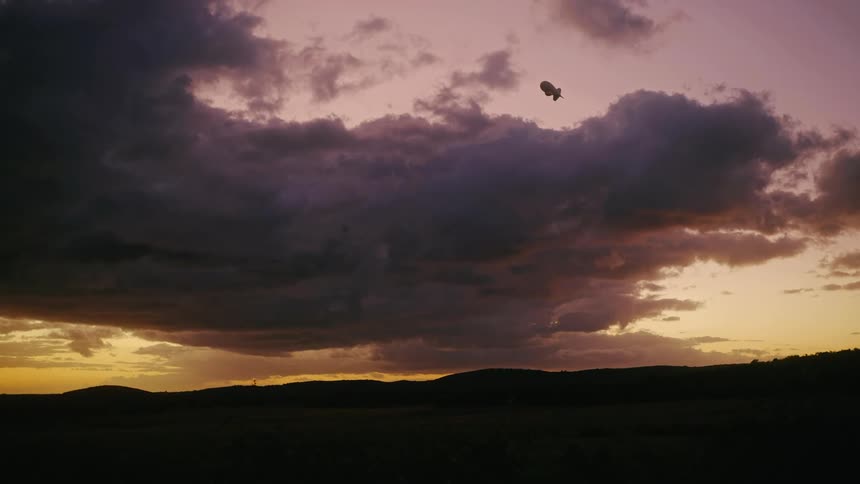 Against a dramatic sunset sky with dark heavy clouds a single airship floats above a dark silhouetted landscape
