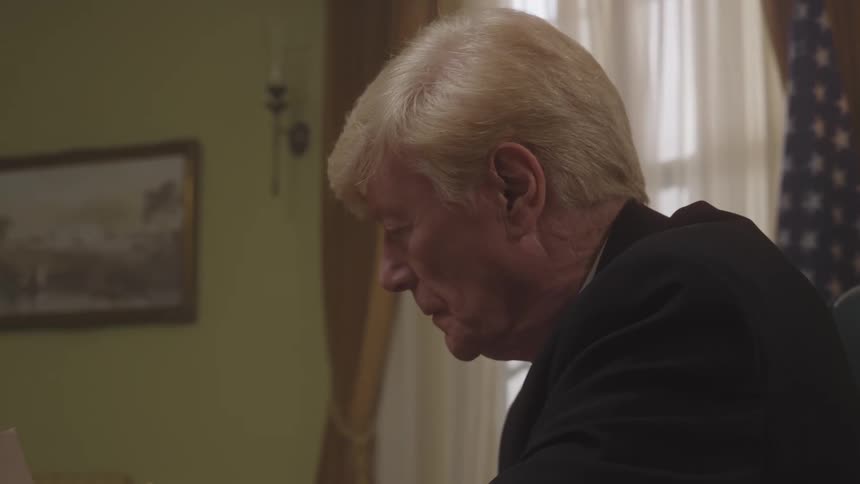 The face of an elderly man in a black suit who is writing something on paper and then turns the paper forward
