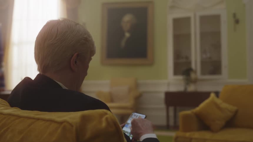 The back of the head of a grayhaired man who is using his cell phone while sitting on a yellow sofa