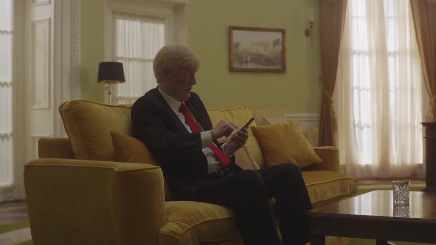 A man with gray hair and wearing a suit with a red tie is sitting on a yellow sofa using his cell phone
