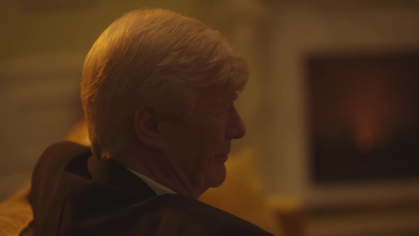 The face of a grayhaired man who is smiling looking forward and wearing a black suit in a room with yellowish lighting