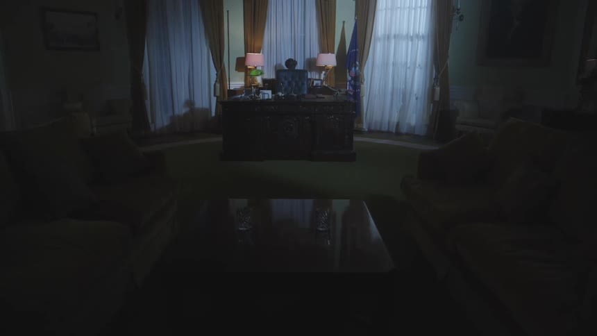 A dimly lit office features a central desk with two lamps heavy curtains framing large windows and flags