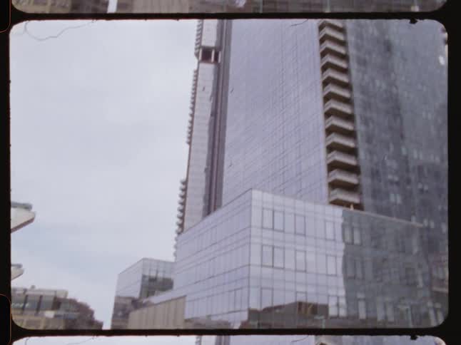An urban train station platform extends beneath towering glass skyscrapers with pedestrians scattered along its edges