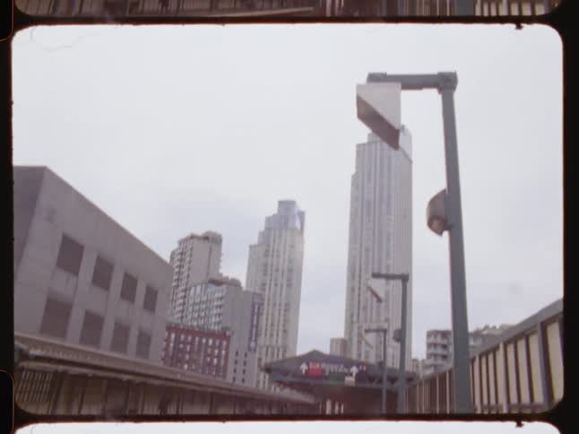A city train station platform with tracks running through the middle surrounded by tall buildings under a cloudy sky