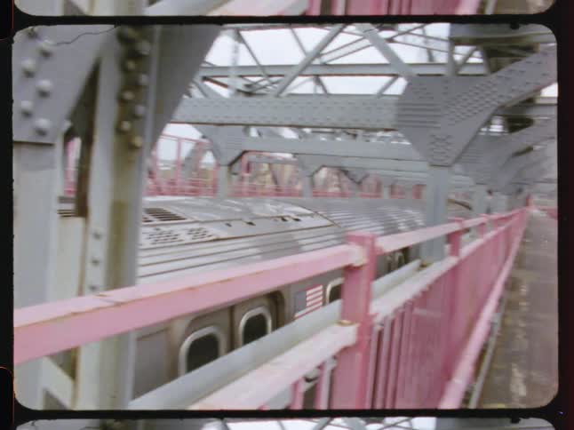 Viewed from an adjacent walkway a silver subway train travels across a large metal bridge structure in older filmstyle footage