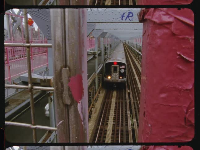 An elevated subway train approaches on tracks running along a metal bridge viewed through the structures pillars and fencing in archival footage