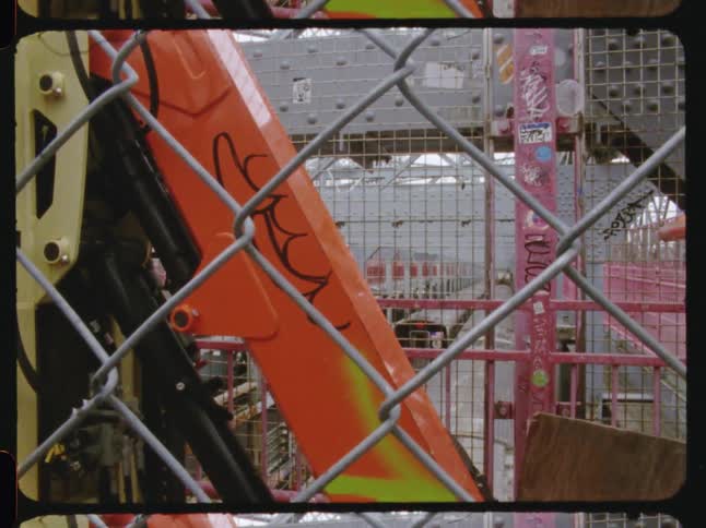 Surrounded by metal fencing orange construction equipment with graffiti appears on an urban site with trains passing in the background