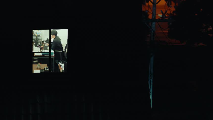 Person working in a bright office seen through a window at night contrasting with the dark outside environment