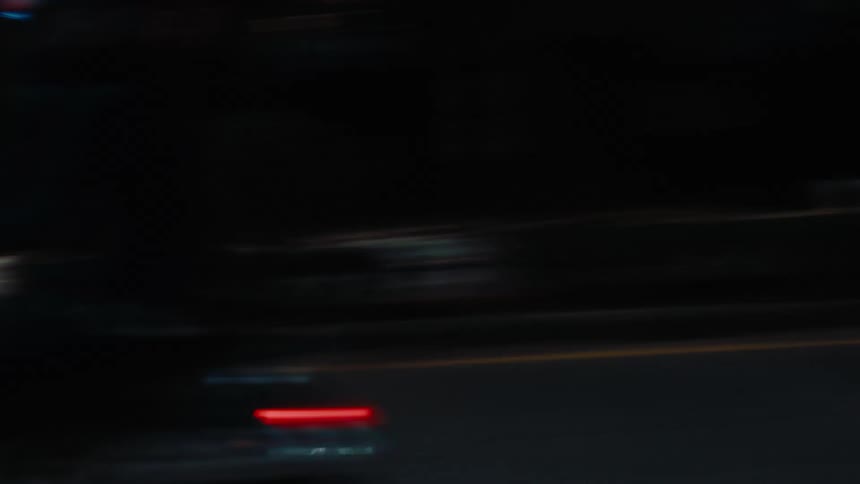 A person riding a scooter at high speed on a city road at night wearing a red helmet with cars passing by in the opposite lane