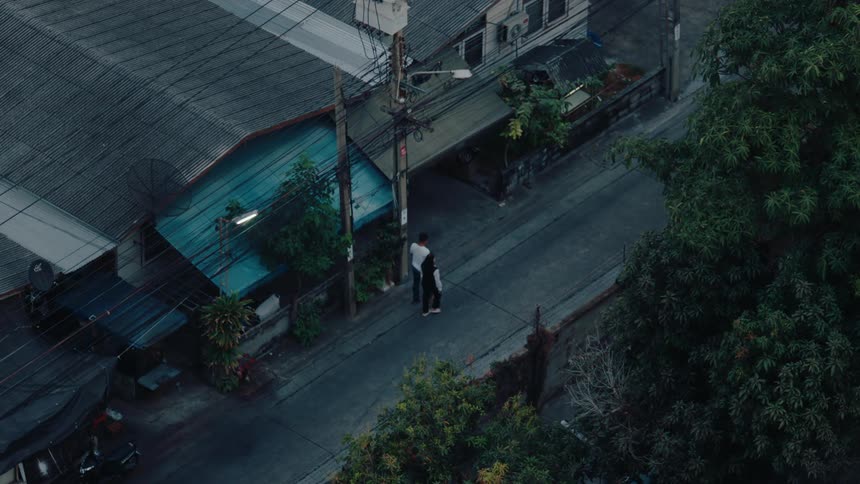 A narrow street with dense houses trees overhead power lines pedestrians and motorcycles