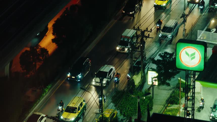 A series of motorcycles driving on a busy road alongside a line of cars all with their headlights on due to nighttime with utility poles trees and plants lining the road