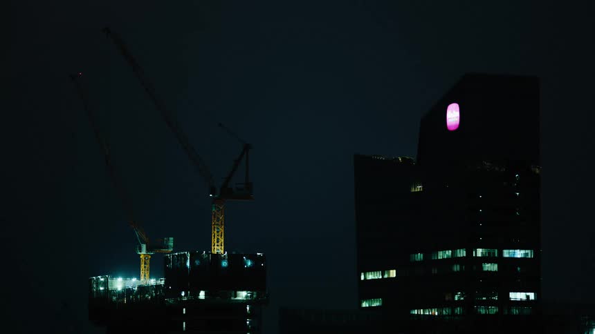 A dark city skyline with a series of lights shining from inside buildings and a blinking light visible at the edge of a crane in a construction site at night